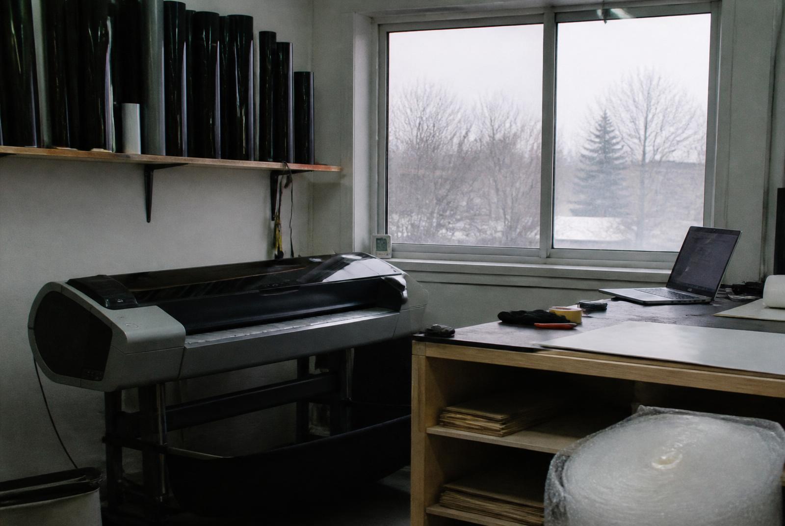 A small Canadian workshop with a cutting plotter, rolls of dark ceramic film, and a winter window.